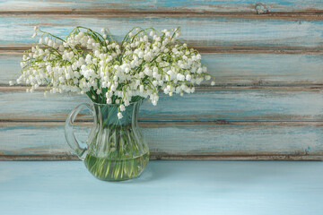 A beautiful bouquet of lilies of the valley on a blue background. White flowers in a glass jug. Wallpaper, greeting card, template. There is a place for text
