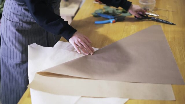 Close up of an unrecognizable woman working in a flower shop cutting and folding paper on desk to wrap flowers. Handheld real time close up shot