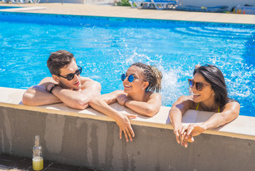 Party at smimming pool. Group of cheerful friends at the edge of the swimming pool drinking cocktails and talking and smiling
