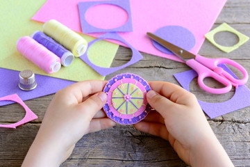 Obraz premium Small child made a simple and colorful flower from felt circles and beads. Child holds a bright felt flower in hands. Craft supplies on wooden table. Easy and fun teaching small kids to sew by hand