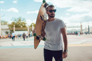Stylish man in sunglasses and with a beard stands on the street with a long board. T-shirt mock up. - obrazy, fototapety, plakaty