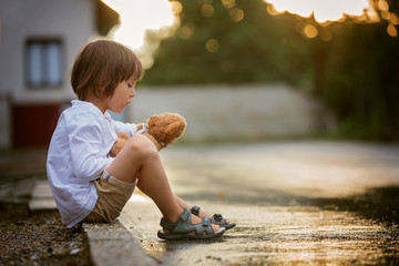 Sad little boy, sitting on the street in the rain, hugging his teddy bear