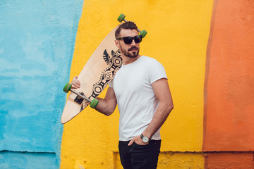 Stylish man in sunglasses and with a beard stands on the background of a multi-colored wall with a long board. T-shirt mock up. © kanashkin