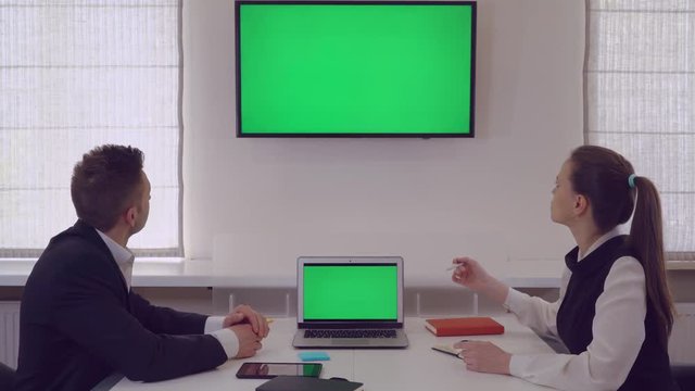 Manager giving presentation for client showing on monitor with green screen. Middle aged businesswoman wearing in elegant black dress. Two people has meeting in modern office.