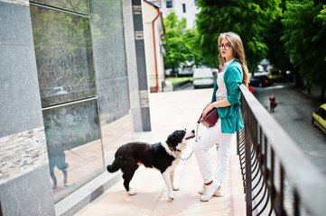 Trendy girl at glasses and ripped jeans with russo-european laika (husky) dog on a leash, against building on street of city. Friend human with animal theme.