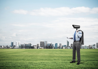 Camera headed man standing on green grass against modern citysca
