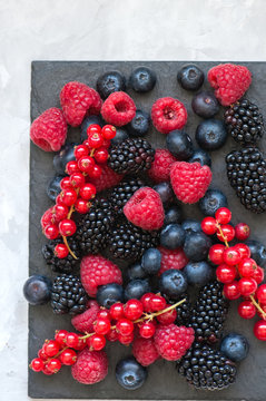 Mix of berries raspberries red currants blueberries and blackberries on black slate board. White stone background.  Overhead view and copy space