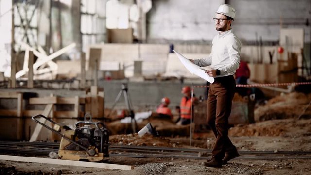 An adult architect considers a construction site where construction is taking place, a person compares the object with the plan and drawings