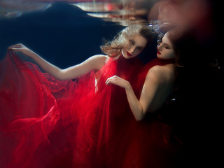Underwater portrait ot two young beautiful girls with make up in red stylish dresses in the swimming pool