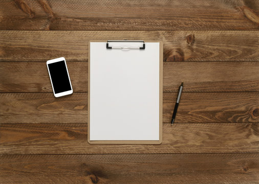 Tabla portadocumentos, smartphone y bol&iacute;grafo sobre fondo marr&oacute;n de madera