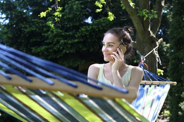 Relaks w ogrodzi.Kobieta rozmawia przez telefon siedząc w hamaku - obrazy, fototapety, plakaty