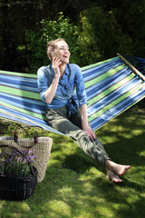 Młoda atrakcyjna kobieta wypoczywa na hamaku w ogrodzie rozmawiając przez telefon - obrazy, fototapety, plakaty