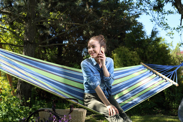 Mój piękny ogród.Młoda atrakcyjna kobieta wypoczywa na hamaku w ogrodzie rozmawiając przez telefon - obrazy, fototapety, plakaty