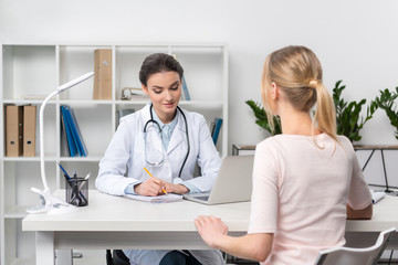 Young doctor with stethoscope taking notes while talking with patient in clinic