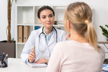 Young doctor with stethoscope taking notes while talking with patient in clinic