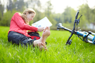 Girl cyclist on a halt reads lying in fresh green grass barefoot