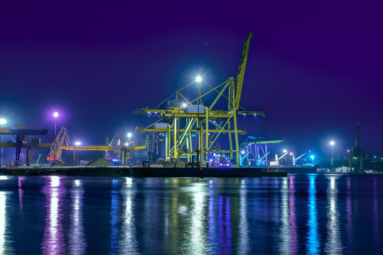 Type of night port. Working freight cranes, a ship at the pier, work in the port. Night photo view of the loading. Beautiful reflection of lights in the sea water.
