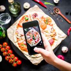 Tasty pizza with ingredients, spices and mobile phone in hand on black background. Flat lay, top view.
