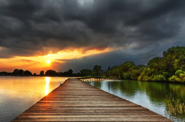 Wooden path bridge over lake at stormy dramatic sunset