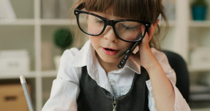 Portrait of caucasian young girl with glasses celling on the cell phone and writing something in notebook, siting on the desk in office. Indoor.