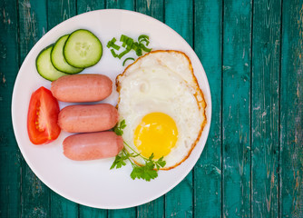 Breakfast fried egg and cooked mini sausages with vegetables on white round plate and on the background of vintage dark turquoise wooden table