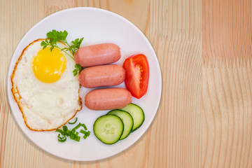 Breakfast fried egg and cooked mini sausages with vegetables on white round plate on light wooden table top view