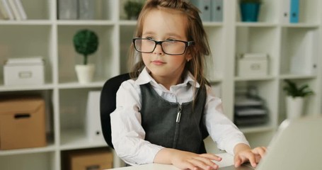 Caucasian pretty young girl with glasses and brown hair typing on the laptop, laying down hands on head, adjusting the glasses on office background. Indoor.