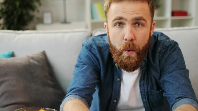Happy young emotional hipster playing game on xbox and drinking coca cola while sitting on sofa at home.