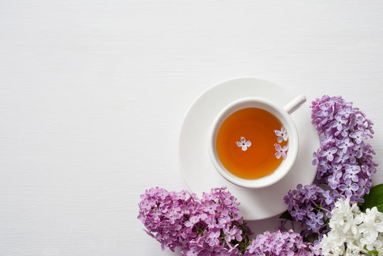 Cup of tea and flowers of a lilac on a white wooden surface, top view