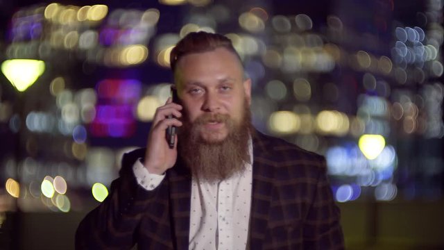 Stylish tourist with a big beard talking on the mobile phone standing in the night city, business center in the background lit by the lights of the Windows of the buildings.