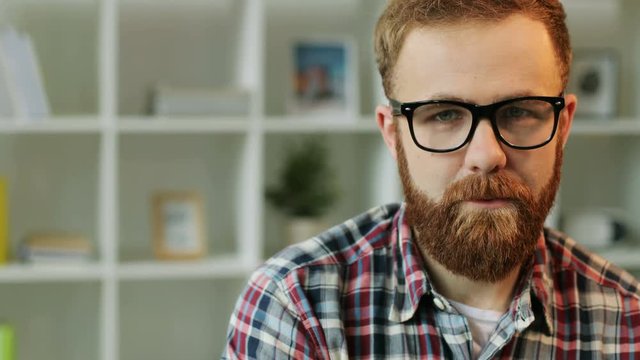 Portrait of serious young man in glasses who start laughting on camera on living room background. Close up.