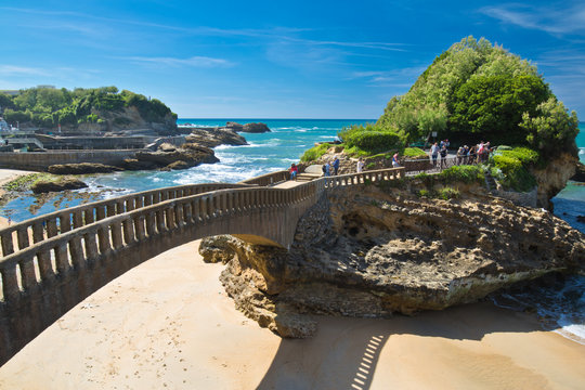 people walking on footbridge leading to cliff island over sandy beach in touristic destination surf spot with turquoise ocean and waves in biarritz in blue sky, basque country, france