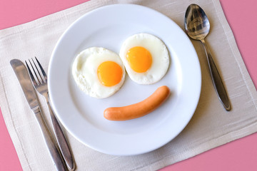 Smiling face frying eggs breakfast on a white plate background with spoon,knife and fork on a fabric table, breakfast serve for kid.