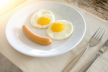 Smiling face frying eggs breakfast on a white plate background with spoon,knife and fork on a fabric table, breakfast serve for kid.