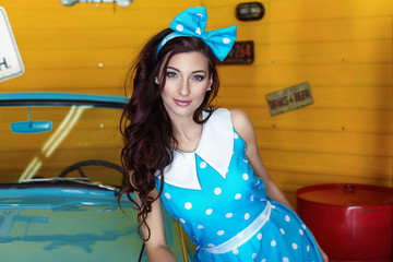 Portrait of a beautiful brunette woman in a blue dress in polka dots with a bow on her head standing near a blue car