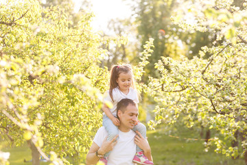 Obraz premium Attractive young man is playing with his daughter in the nature. The father is standing and carrying girl on his back. He is stretching arms sideways. The family is smiling
