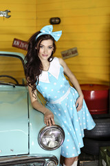 Young beautiful brunette woman dressed in a blue dress with polka dots with a bow on her head stands leaning on a blue car