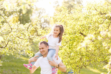 Attractive young man is playing with his daughter in the nature. The father is standing and carrying girl on his back. He is stretching arms sideways. The family is smiling
