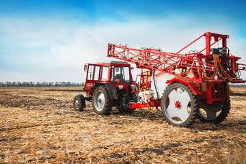Red tractor with trailed sprayer in a field against a blue sky.