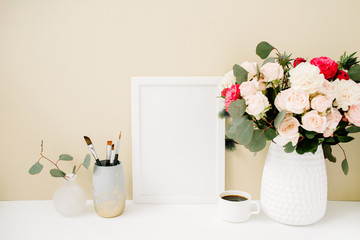 Home office desk with photo frame mock up, beautiful roses and eucalyptus bouquet in front of pale pastel beige background. Blog, website or social media concept .