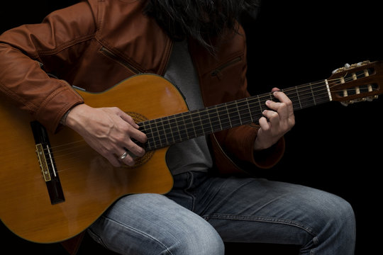 Long hair male guitarist playing with acoustic guitar in leather jacket on the stage.