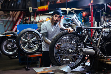 Portrait of bearded  tattooed man working in garage tuning  motorcycle and looking at camera - obrazy, fototapety, plakaty
