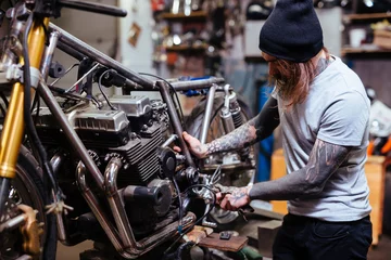 Photo sur Plexiglas Moto Portrait d& 39 un homme tatoué concentré travaillant dans un garage personnalisant une moto et réparant des pièces cassées