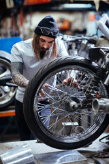 Portrait of modern  tattooed man working in garage customizing  motorcycle and repairing broken wheel mechanism