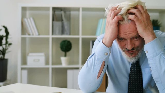 Caucasian midle aged man sitting on the desk holding his head of hands in office background. Indoor. Close up.