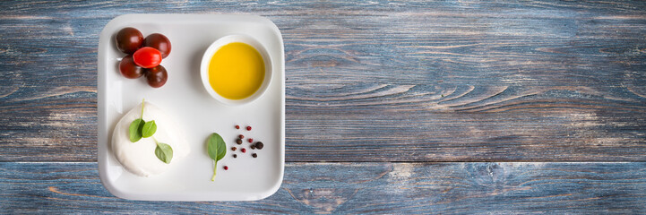 Pickled cheese mozzarella with olive oil, cherry tomatoes, green basil leaves and spice peppercorns on white plate on rustic wooden table. Top view. Wide panoramic image. Copy space.