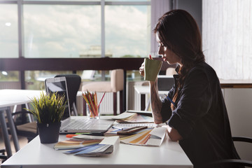 Portrait of beautiful happy  young designer woman holding cup of tea while  sitting at the desk with instruments, colour samples and laptop.