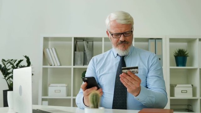 Caucasian midle aged business man typing credit card numbers on smart phone for online shopping on office background. Indoor.