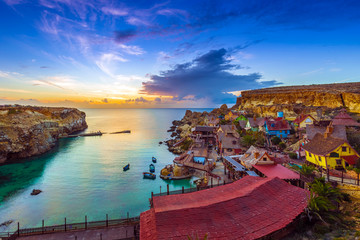 Mellieha, Malta - Skyline view of the beautiful Popeye Village at Anchor Bay at sunset with amazing colorful clouds and sky - obrazy, fototapety, plakaty