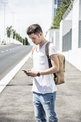 Handsome trendy young man wearing white shirt standing and using cell phone, outdoor in city setting in day shot on sidewalk by street